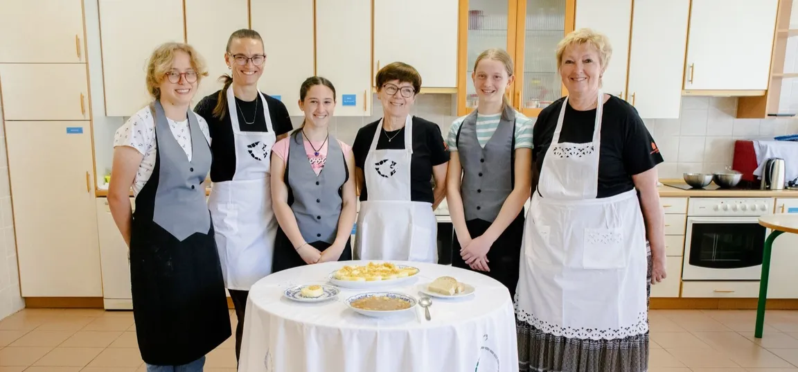 Students and their mentors, dressed in aprons, stand in the classroom. In front of them on a round table covered with a white tablecloth are typical Notranjska dishes - kavra and cottage cheese štruklji.