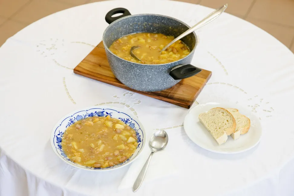 On a round table covered with a white tablecloth, stands a kavra (kohlrabi stew) in a white soup plate with a blue pattern. Next to it are two slices of bread and a kavra container with a ladle.