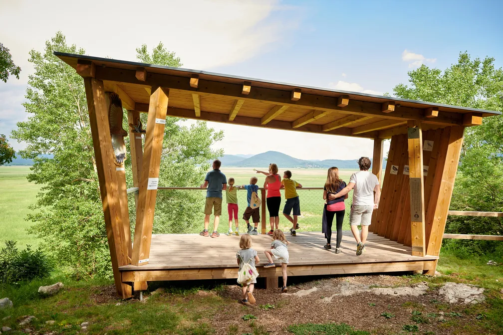 Skupina ljudi na opazovalnici občuduje razgled na polje. Obdaja jih zeleno drevje, nad njimi je jasno nebo.