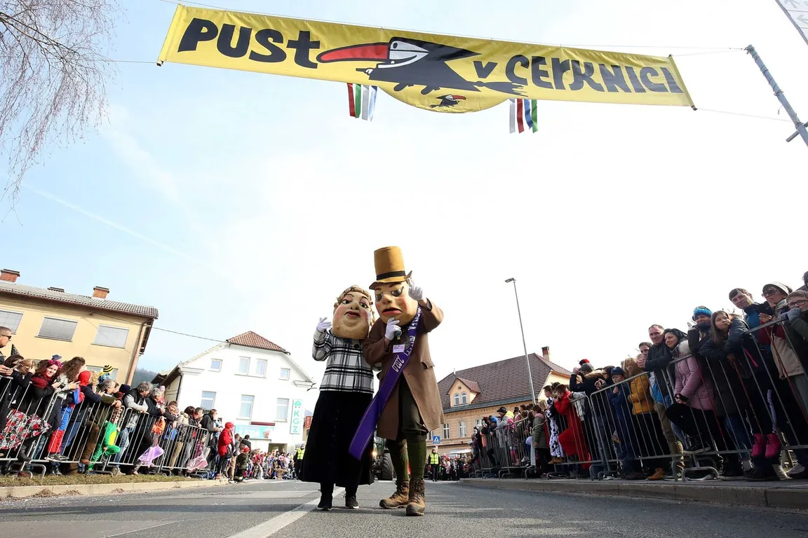 Dve osebi v kostumih stojita po rumenim napisom "Pust v Cerknici", množica ljudi stoji za ograjami. Nad njimi je jasno nebo.