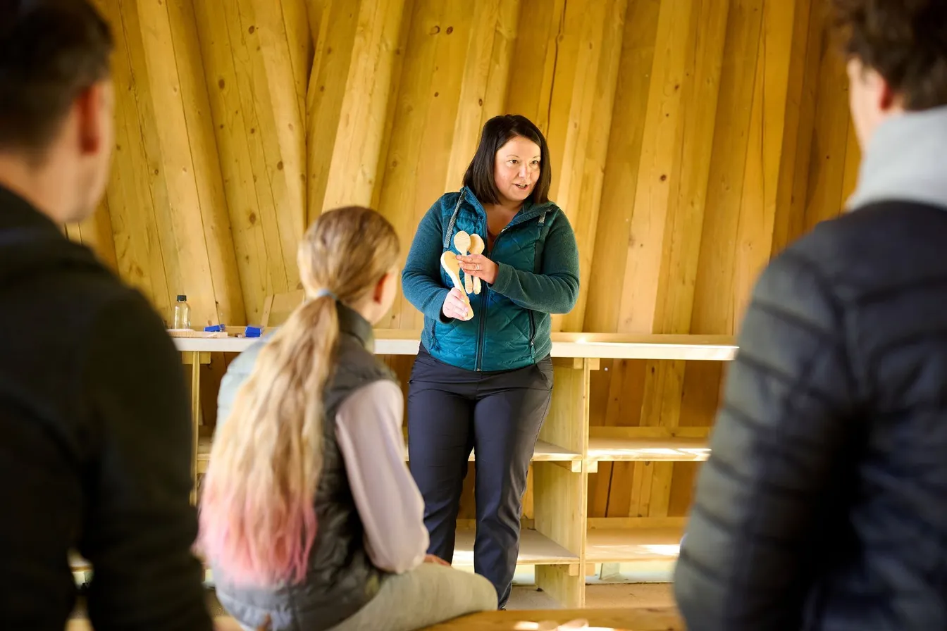 A guide presents a carving activity in the forest. She is standing in the outdoor classroom "Hlejvi" by Lake Cerknica, holding three hand-carved wooden spoons in her hands. In the foreground, three people are sitting on benches and listening to her.