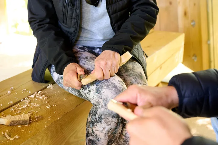 Two people shape wooden spoons with special knives for shaping wood products. They sit on benches in an outdoor classroom, with wooden chips on the benches next to them.