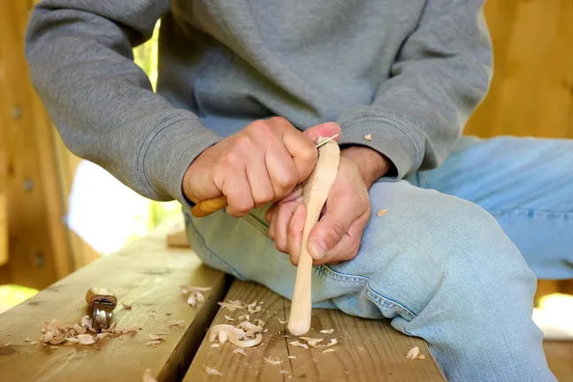 A person is carving a wooden spoon by hand. He is sitting on a wooden bench in an outdoor classroom, next to him there are shavings and knives intended for shaping wooden products.