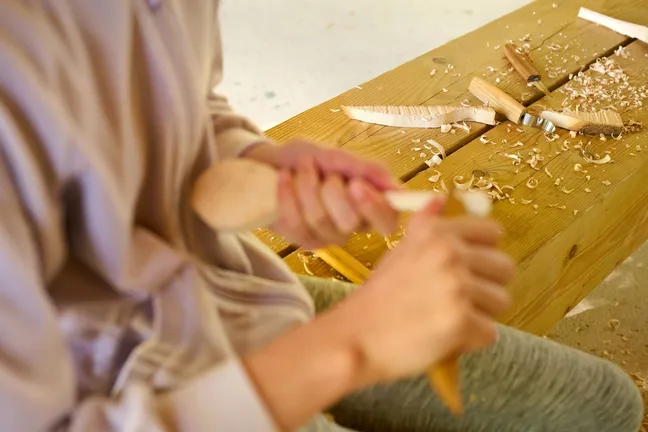 A person is carving a wooden spoon by hand. In the background, on the wooden bench on which he is sitting, there are visible shavings and various knives and blades intended for shaping wooden products.