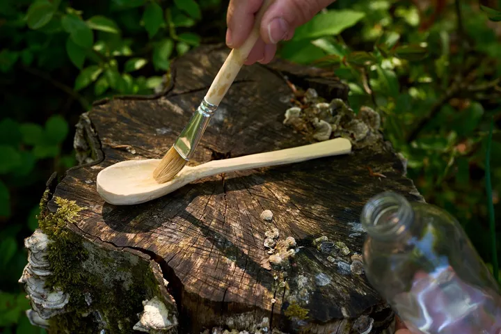 A hand holds a brush and paints a wooden hand-carved spoon with a transparent coating - oil. The spoon is placed on an old wooden stump in the forest, surrounded by green leaves.