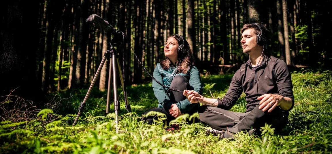 A couple sits on the forest floor surrounded by lush greenery and tall trees, listening to nature sounds through headphones with their eyes closed. A microphone on a tripod is placed in front of them, pointed into the forest.