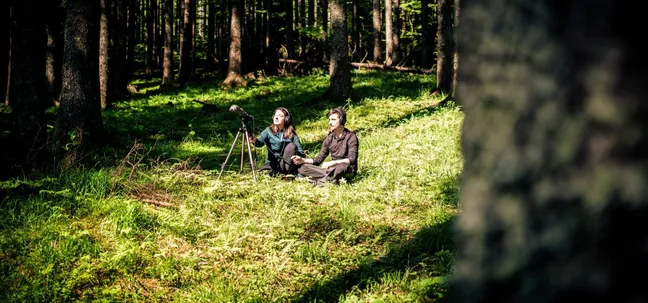 A wide view of a couple sitting on soft forest grass in a bright clearing surrounded by tall trees, listening to nature sounds through headphones, with a microphone on a tripod set up in front of them.