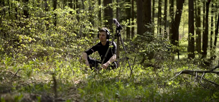 Close-up of a microphone on a tripod in the foreground, with a man wearing headphones sitting in the forest in the background, slightly out of focus as he listens to nature sounds.