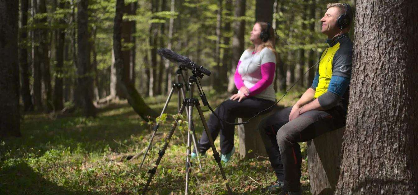 A man and a woman sit on a wooden bench in the forest, relaxed and listening to nature sounds through headphones, with a microphone on a tripod set up in front of them.