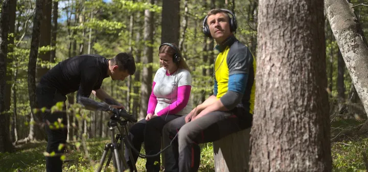 Three adults take part in an audio experience in the forest; two sit on a bench listening to nature sounds through headphones, while the third adjusts recording equipment with a microphone on a tripod.
