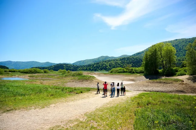 Birdwatchers stand by the dried-up Lake Cerknica. They are equipped with binoculars and spotting scopes, in the background the dry lake bed and some water in the riverbed and the Rešeto pond can be seen.