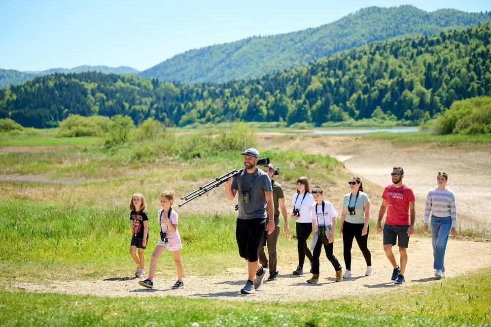 People, adults and children, walk along a dry Lake Cerknica. They are equipped with binoculars and bird-watching spotting scopes, with the dry lakebed visible in the background, and a forest and some water in the Rešeto pond in the far distance.