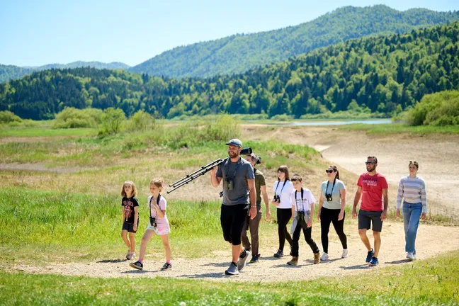 Ljudje, odrasli in otroci, hodijo ob presahlem Cerkniškem jezeru. Opremljeni so z daljnogledi in spektivi za opazovanje ptic, v ozadju je videti suho jezersko dno, čisto zadaj pa gozd in nekaj vode v ribniku Rešeto.