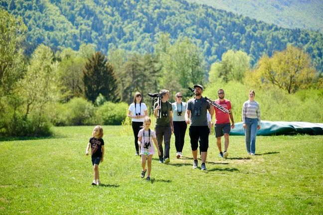People, adults and children, walk along a meadow by Lake Cerknica. They are equipped with binoculars and bird-watching spotting scopes and there are canoes in the background.