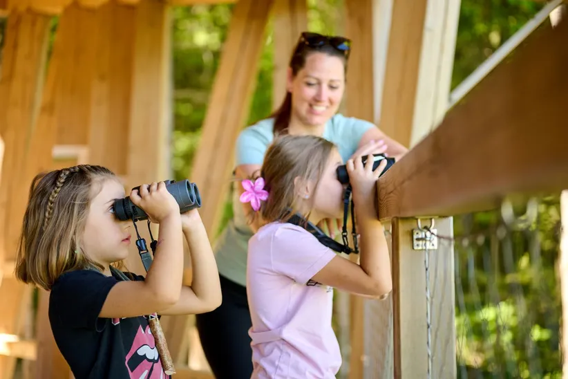 Two girls are watching birds on Lake Cerknica with binoculars. In the background is a smiling woman. They are standing at the Kuharca observation platform on the Drvošec trail.