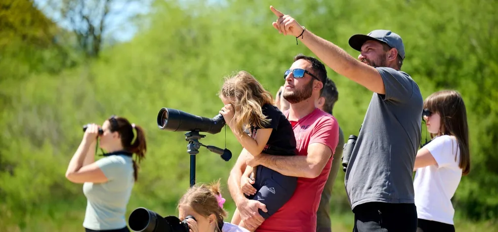 A group of birdwatchers stands by Lake Cerknica. Adults and children with binoculars and bird-watching telescopes gaze into the distance. A guide wearing a hat points to the sky with his finger.
