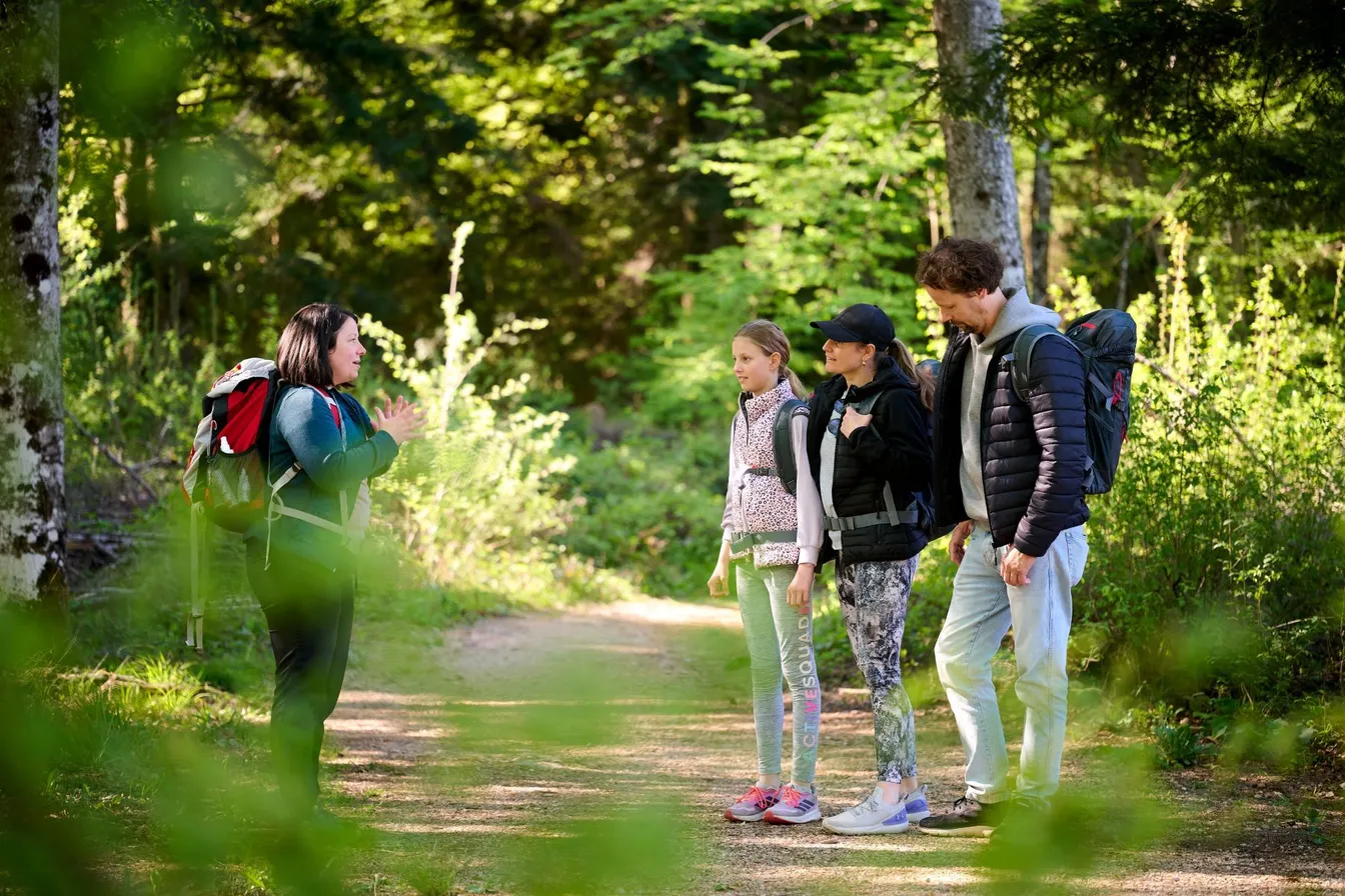 A guide with a backpack is describing the "Hidden Sounds of the Forest" experience to three participants. They are standing on a forest path, surrounded by green bushes and trees.