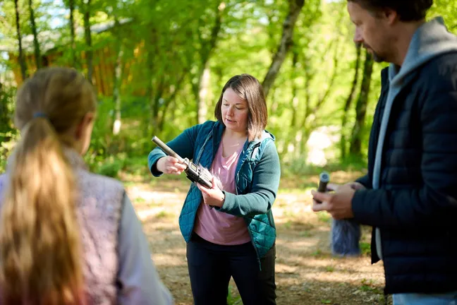 A guide in the forest is holding a special microphone for capturing the sounds of the forest and is explaining how the microphone works to the people standing next to her.