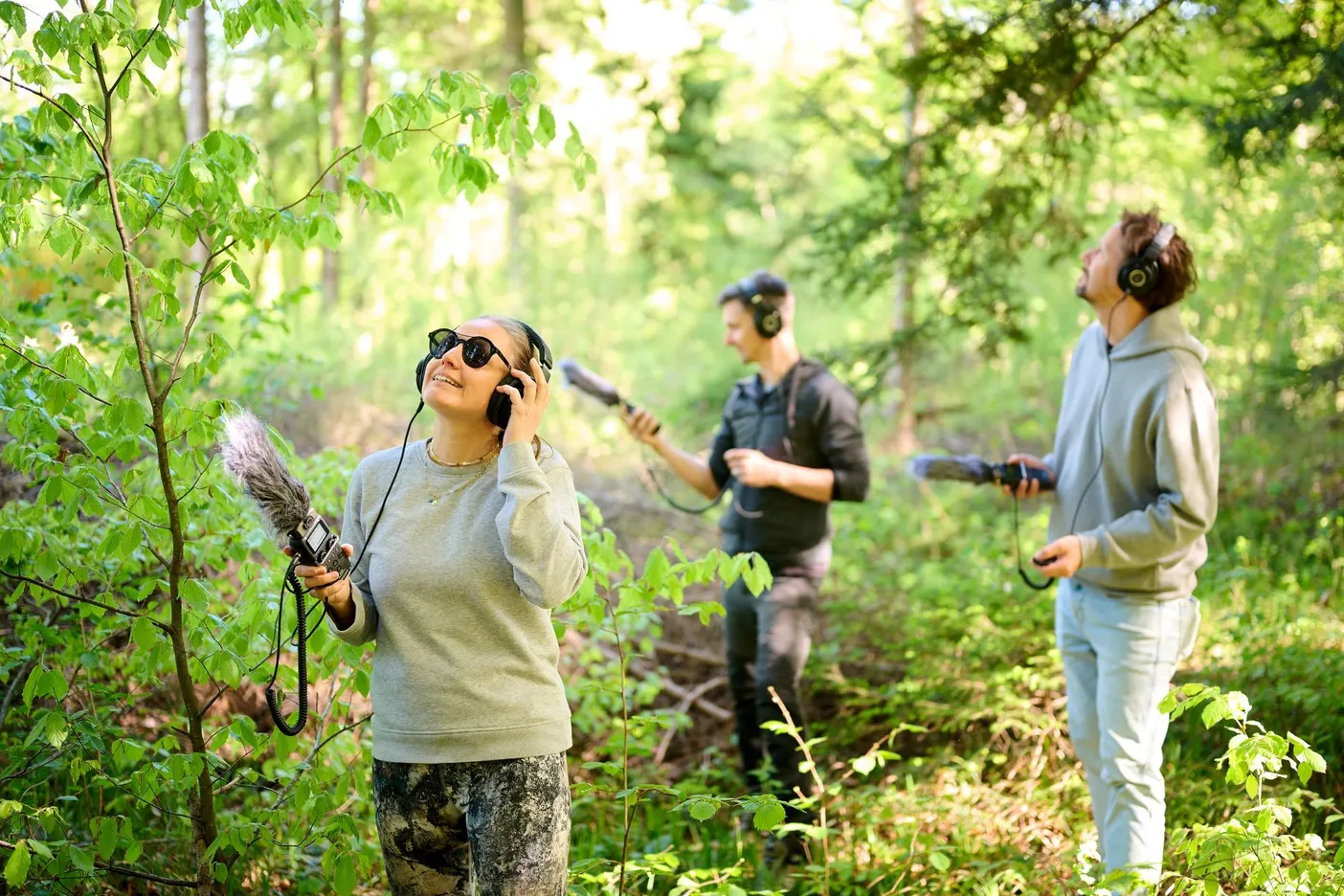 Three people are standing in the forest, equipped with various microphones and headphones. They look around with interest and listen to the sounds of the forest around Lake Cerknica.