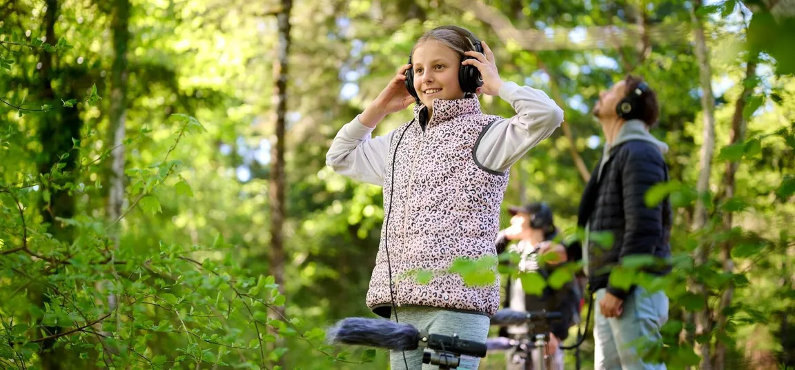 Deklica nasmejano stoji v gozdu s slušalkami na ušesih. Pred njo je mikrofon, s pomočjo katerega posluša zvoke gozda. V ozadju za njo sta še dve odrasli osebi, ki prav tako vsaka s svojim mikrofonom in slušalkami poslušata dogajanje v okolici.
