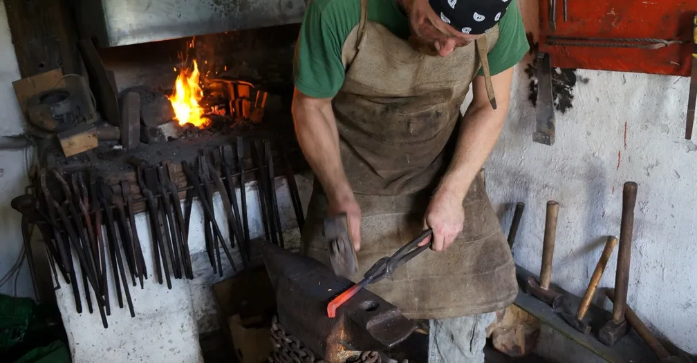 A blacksmith at work in his workshop, holding red-hot iron with tongs and striking it with a hammer.