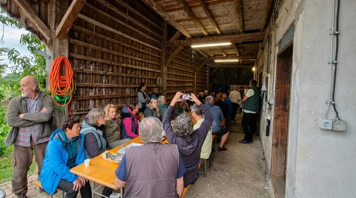 Udeleženci dogodka sedijo za dolgimi mizami v odprtem lesenem gospodarskem poslopju in spremljajo predstavitev. V ospredju je več oseb na klopeh, ena fotografira dogajanje s telefonom. Na steni visi zvita vrtna cev, prostor pa osvetljujejo stropne luči. V ozadju je zbrana skupina poslušalcev.