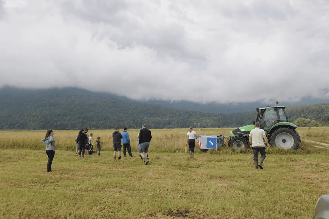 The picture shows a group of people standing on a meadow, with a tractor next to them. The sky is cloudy, and mountains can be seen in the background.