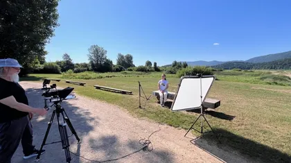 Oseba sedi na klopi sredi travnika, obkrožena s snemalno opremo, vključno s kamero na stojalu, osvetlitvijo in reflektorjem; v ozadju drevesa, hribi in jasno modro nebo.