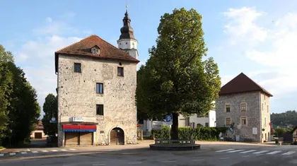 Obrambna stolpa z rdečo streho na križišču, v ozadju zvonik cerkve. V središču križišča stoji veliko drevo, obdano s krožno klopjo. Nebo je jasno z nekaj oblaki, brez vidnih ljudi ali vozil