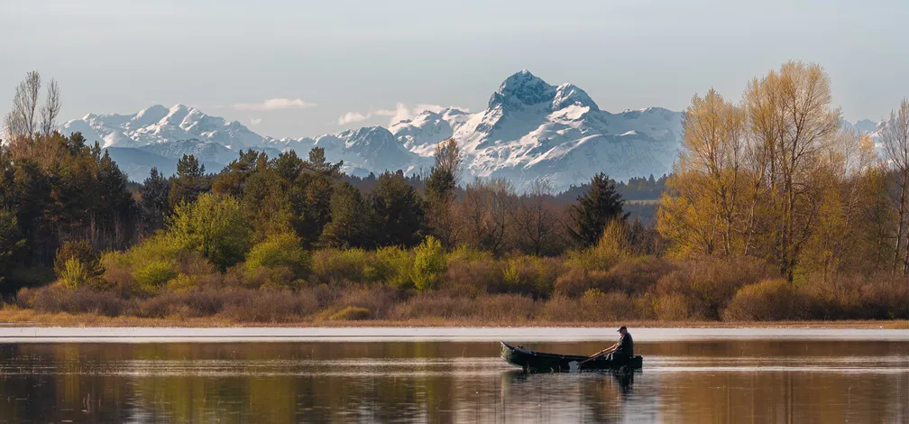 Oseba vesla v čolnu na mirnem jezeru, obdanem z drevesi v jesenskih barvah, v ozadju pa se dvigajo zasneženi gorski vrhovi pod jasnim nebom.