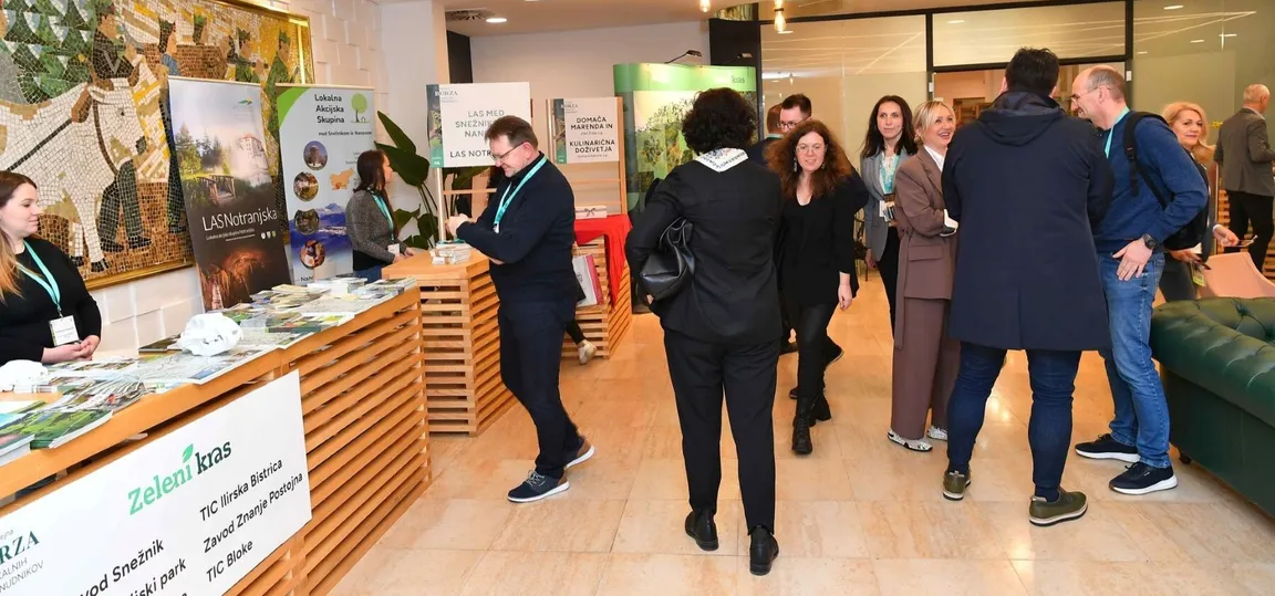 Notranji razstavni prostor z več promocijskimi stojnicami turističnih organizacij, kjer se obiskovalci pogovarjajo in pregledujejo brošure.