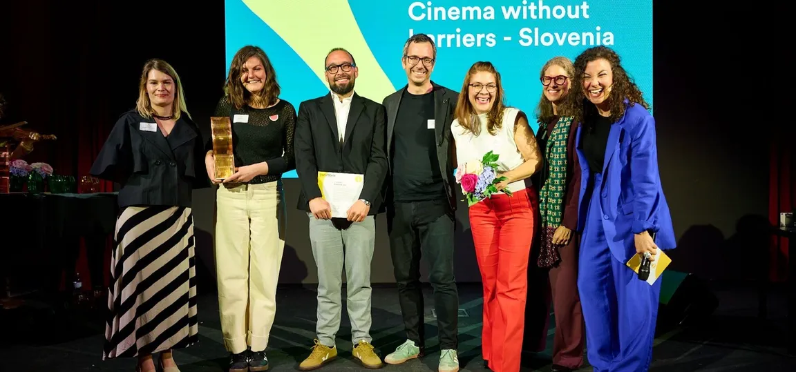 Skupina predstavnikov projekta »Cinema without Barriers – Slovenia« stoji na odru ob prevzemu 3. nagrade SozialMarie. Udeleženci se smejijo in pozirajo pred projekcijskim zaslonom z imenom projekta, ena izmed nagrajenk pa drži bronast kip nagrade.