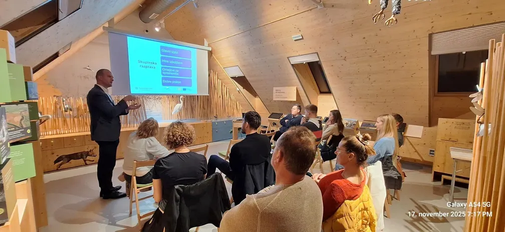 Presenter explains the hospitality module as part of the NRP Academy project (LAS Notranjska), while participants sit in a wooden meeting room watching the projection.