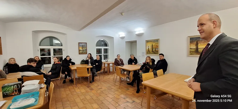 Skupina ljudi sedi za lesenimi mizami v svetli sobi z belimi stenami in slikami na stenah. Na levi strani so okna z zaobljenim zgornjim delom, na mizah so krožniki in pladnji. V ospredju stoji oseba v temni obleki, prostor ima tlakovano rjavo talno oblogo.