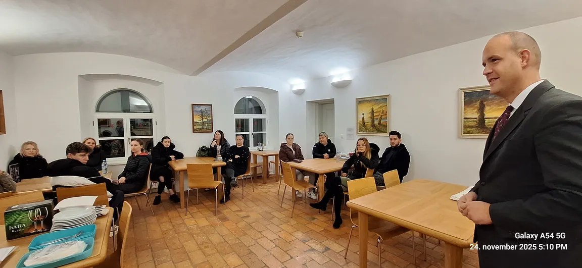 Skupina ljudi sedi za lesenimi mizami v svetli sobi z belimi stenami in slikami na stenah. Na levi strani so okna z zaobljenim zgornjim delom, na mizah so krožniki in pladnji. V ospredju stoji oseba v temni obleki, prostor ima tlakovano rjavo talno oblogo.