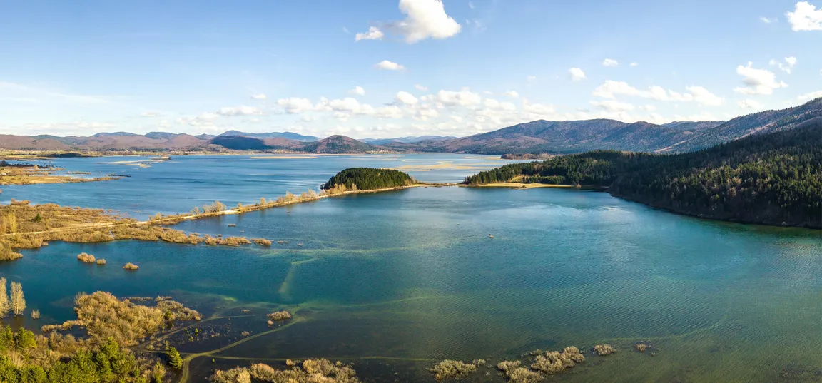 Panoramski pogled na jezero, obkroženo z gozdnatimi griči in hribovji v ozadju pod modrim nebom z nekaj belimi oblaki.