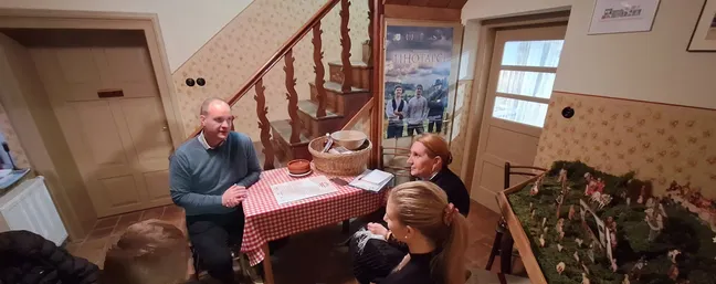 Notranjost domačega prostora z lesenim stopniščem in mizico, prekrito z rdeče-belim karirastim prtom. Na mizi so košara in nekaj papirjev. Ob mizi sedi več oseb, v ozadju so vrata, stena z vzorcem in jaslice na desni strani.