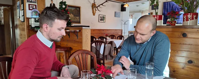 Dva človeka sedita za mizo v rustikalni gostilni, na mizi so kozarci, prtiček in dekoracija z rdečimi rožami. V ozadju so leseni stoli, mize s pogrinjki, lesena stena in okrasni predmeti, vključno z jelenovimi rogovi na steni.