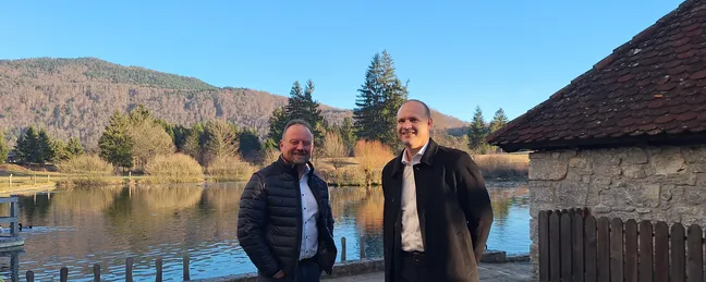 Dve osebi stojita zunaj ob jezeru, v ozadju so drevesa in gozdnati hribi, na desni strani je kamnita stavba z rdečo streho, nebo je jasno modro.