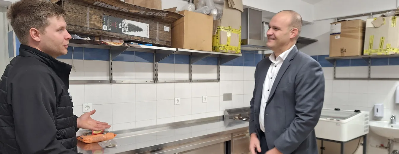 Dva človeka stojita v kuhinji, na steni so police z različnimi kartonskimi škatlami in kuhinjskimi pripomočki.