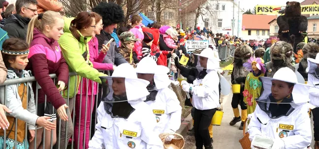 Pustna povorka z liki v kostumih čebel in čebelarjev, medtem ko udeleženci delijo promocijski material obiskovalcem ob cesti.