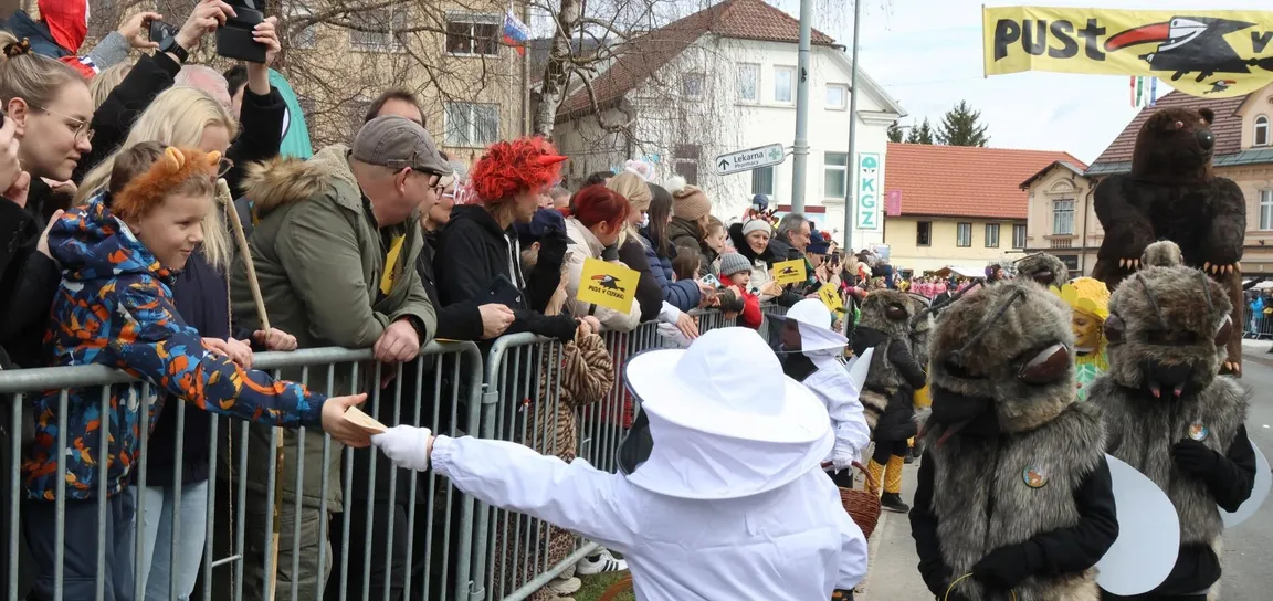 Udeleženec v čebelarskem kostumu med pustno povorko deli promocijski material obiskovalcem za ograjo.