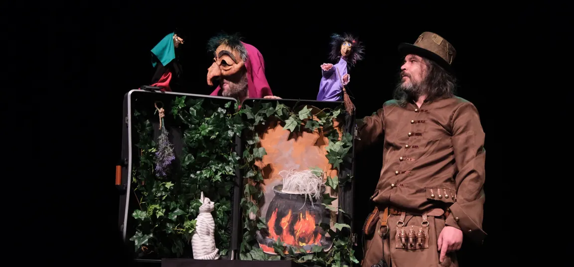 A puppet show on stage. Against a set decorated with greenery and an illustration of a cauldron over a fire, the puppets perform, whilst a puppeteers in costume stands beside them. The dark backdrop emphasises the illuminated action on stage.