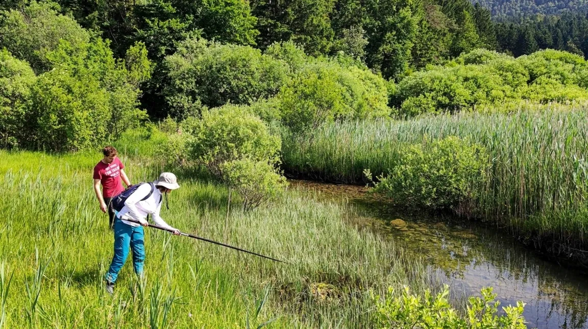 Dve osebi stojita ob majhnem vodnem telesu v zelenem, gosto poraščenem okolju; ena oseba z belim klobukom in modrimi hlačami drži dolgo palico, druga v rdeči majici stoji zadaj.