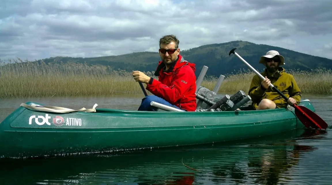 Two people in a green canoe on the water, wearing red and yellow jackets, with oars in their hands; in the background, reeds, hills, and a cloudy sky.