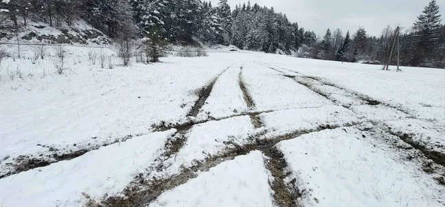 Snežna pokrajina z vidnimi globokimi kolesnicami, ki se križajo na zasneženem travniku. V ozadju so gozdnati hribi, prekriti s snegom, in nekaj dreves ob robu polja. Slika prikazuje poškodovano snežno površino zaradi vožnje vozil po naravi.