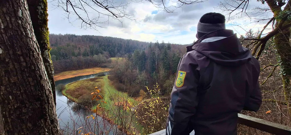 A person wearing a dark jacket with a nature conservation badge on the sleeve stands at a viewpoint looking towards a river winding through a wooded valley. The trees in the foreground are bare, while in the background an autumn landscape stretches out in shades of green and brown.