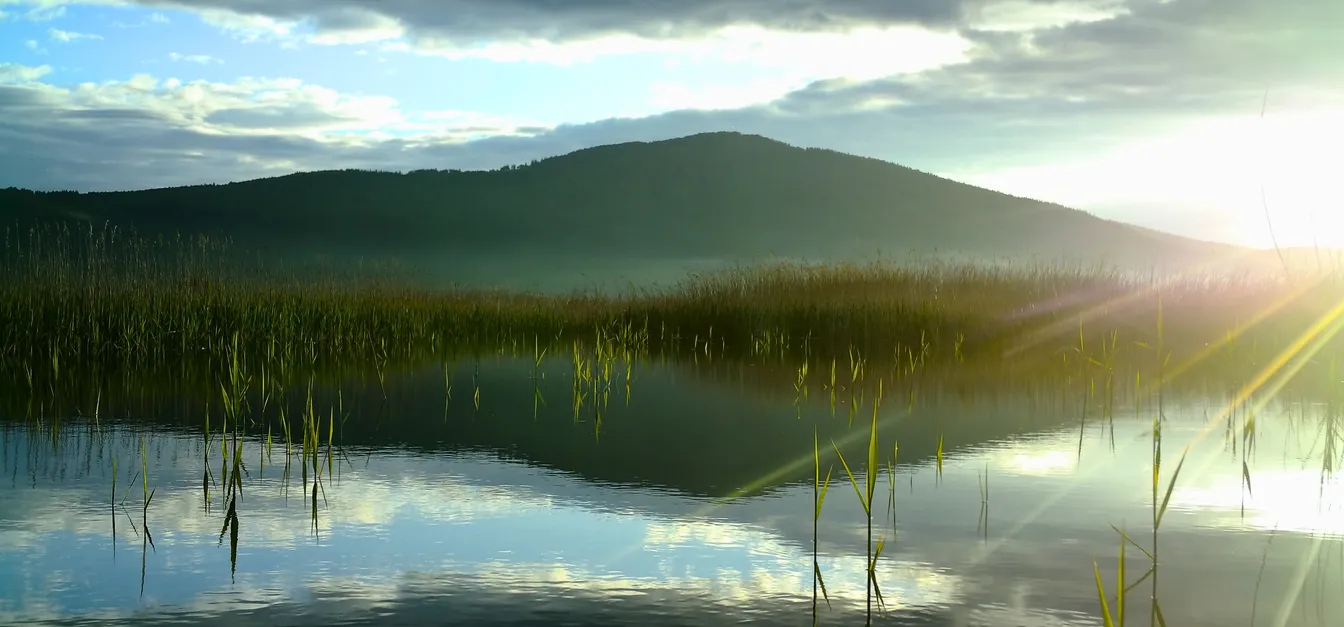 Mirna vodna površina z odsevom neba in oblakov, obdana s trstičjem, v ozadju pa se dviga gozdnat hrib; sončni žarki prodirajo skozi obzorje ob sončnem vzhodu.