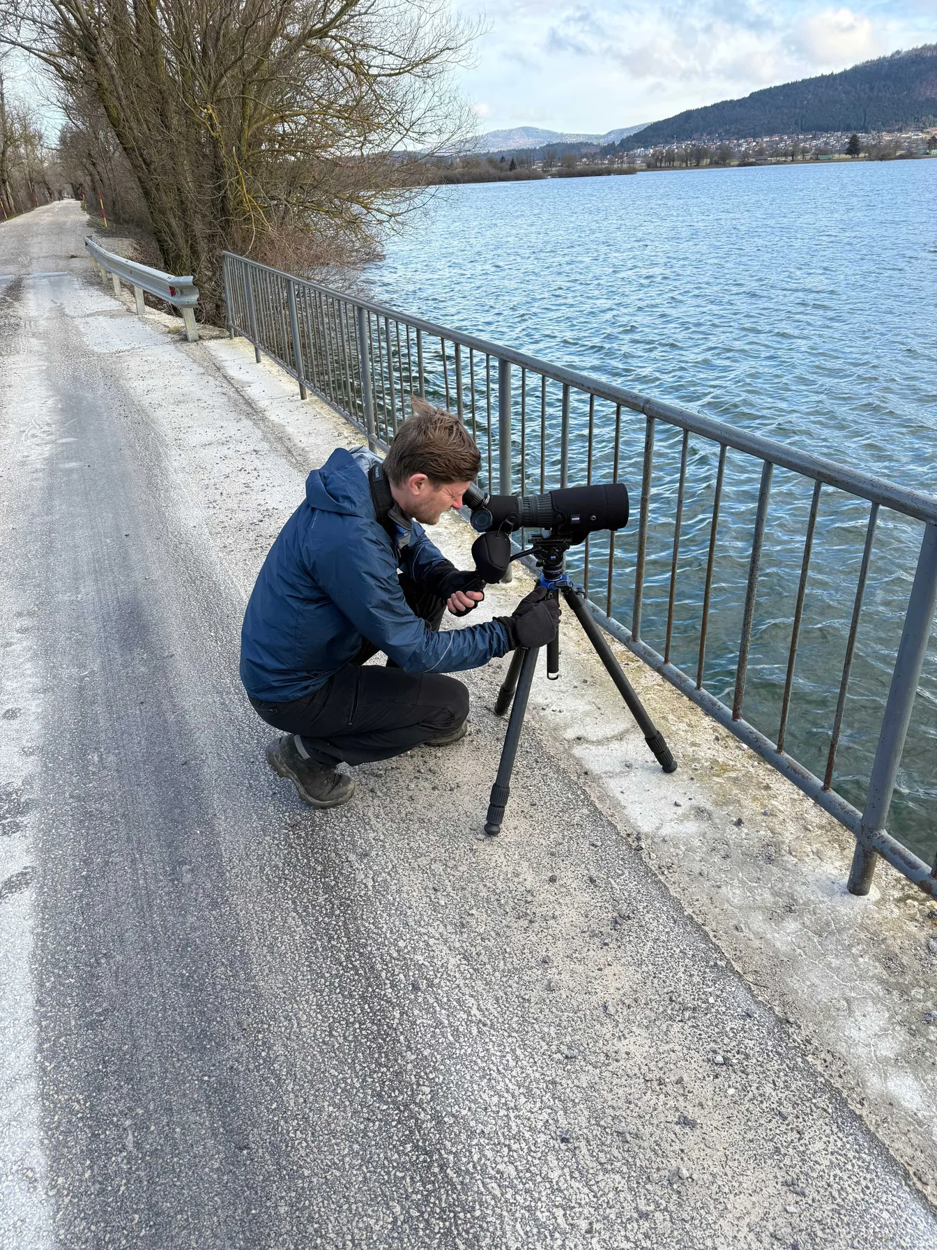 Oseba v modri jakni kleči ob cesti ob vodi in skozi velik teleskop na stojalu opazuje jezero; ob strani je kovinska ograja, v ozadju pa hribovit teren in oblačno nebo.