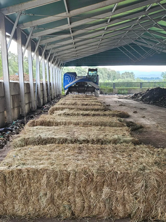 Pokrit kmetijski prostor z odprtimi stranicami, v katerem je na tleh v ravni vrsti zloženih več velikih pravokotnih bal suhe trave ali sena. V ozadju je kmetijski stroj ob večjem kupu rastlinske mase, zadaj pa skozi odprtino objekta posegata zeleno drevje in nebo.